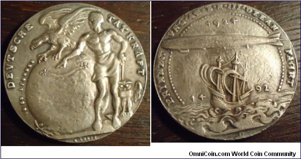 1924 Germany Zeppelin Medal by K. Goetz. Silver: 33MM./18.10 gms.
Obv: Transfer of reparations airship ZR II in the United States. Airship over Columbus caravel. Rev: Scantily clad men in front of globe shape with langgestieltem hammer besdie him. Flying eagle & owl below, flight route is marked on the ball and the flight time given next to it.
